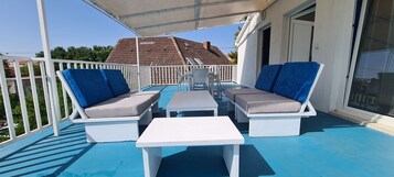 Un espacio de estar al aire libre cubierto con mobiliario blanco y cojines azules, una pequeña mesa y vistas a una casa con techo de tejas.