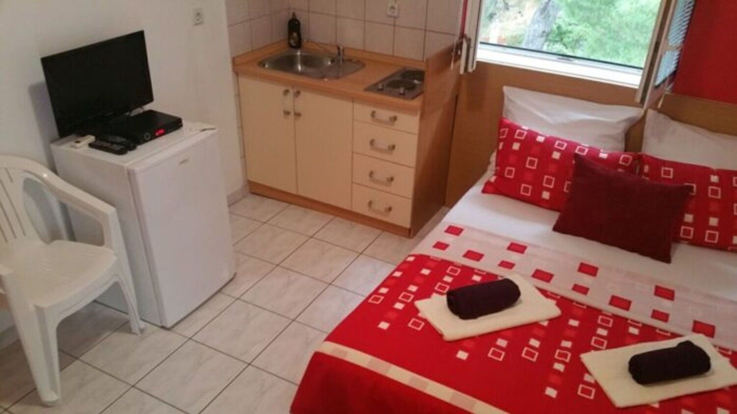 A compact room with a bed, a small kitchenette, a TV, and a chair.