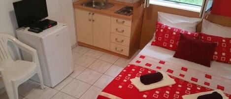 A compact room with a bed, a small kitchenette, a TV, and a chair.