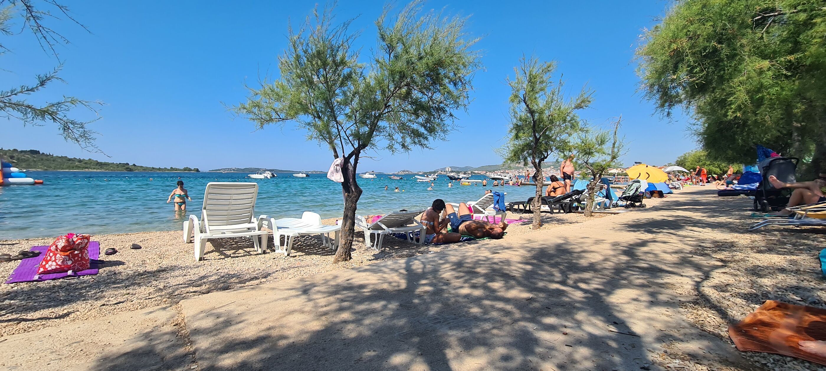 Egy strand, ahol napozóágyak, egy fa és a háttérben csónakok láthatók.