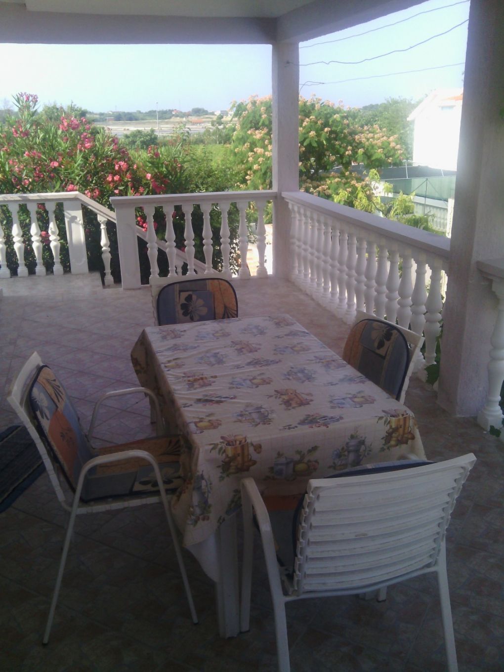 Un área de comedor al aire libre cubierta, con mesa y sillas, rodeada por una barandilla blanca, con vista a un jardín de flores rosadas y vegetación.