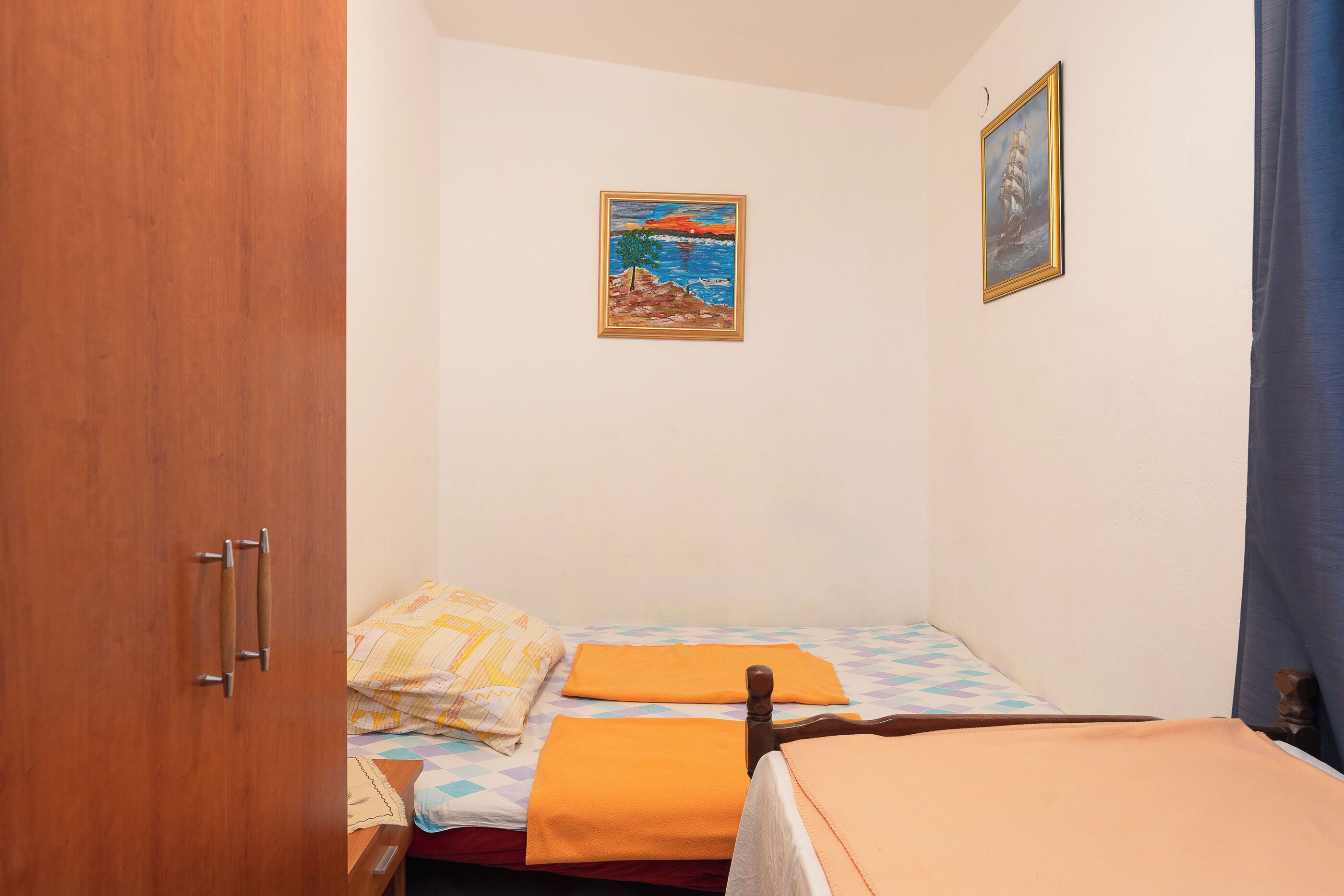 A small bedroom with two beds, a wooden wardrobe, and two paintings on the wall.