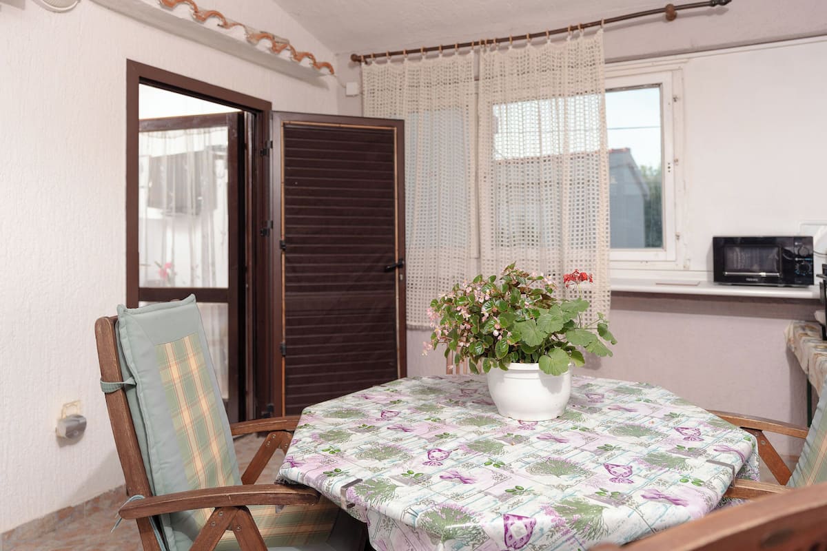 A room with a wooden chair, a table with a floral tablecloth, a potted plant, and a window with curtains.