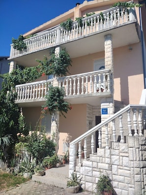 A multi-story building with a stone facade, a balcony with white balustrades, and a staircase leading to the entrance.