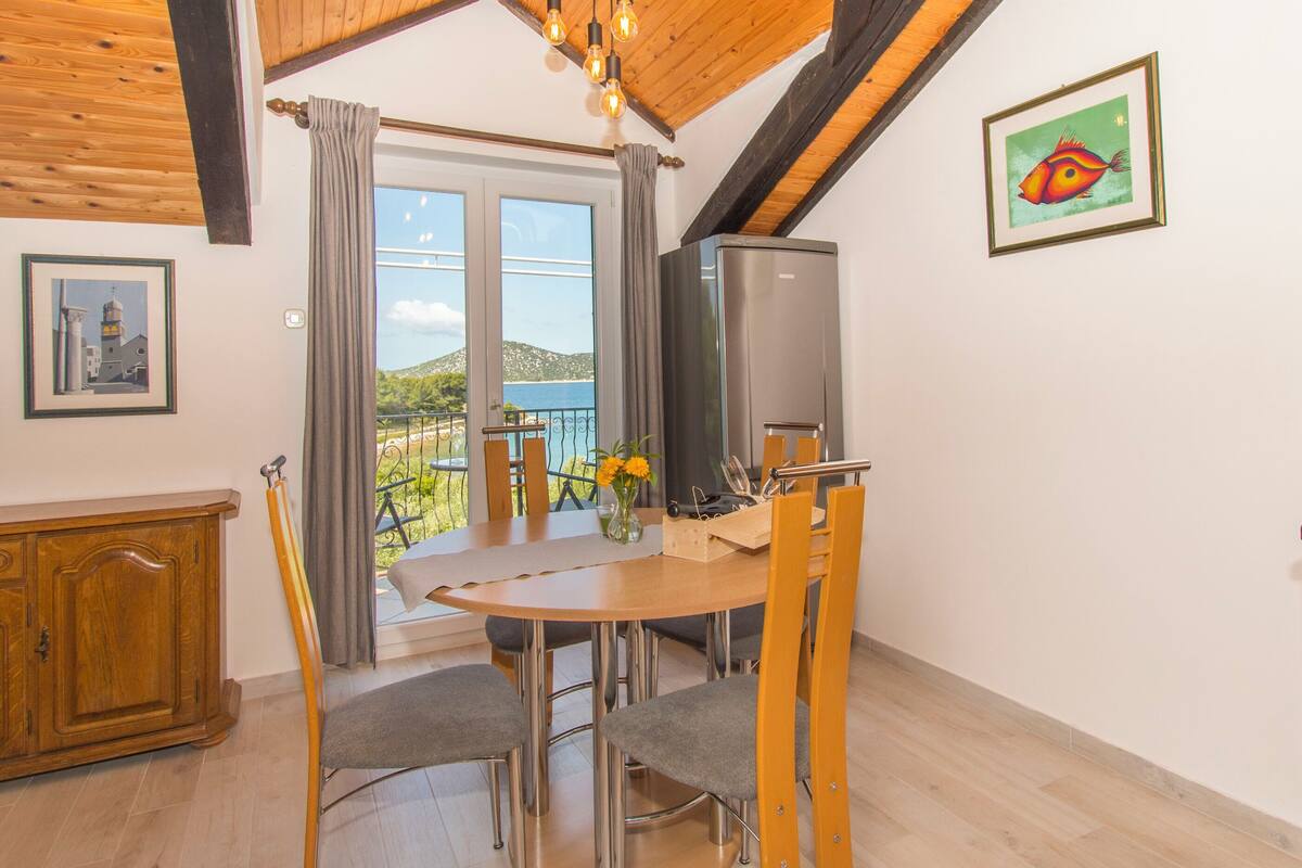 A dining area with a wooden ceiling, a table with chairs, a TV, and a view of the sea through large windows.