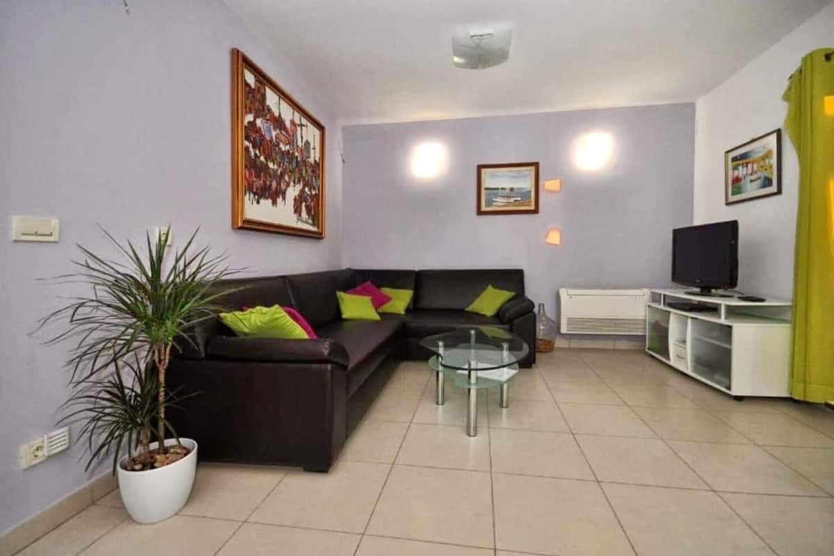A living room with a dark brown sofa, a glass coffee table, a potted plant, a television on a white stand, and a painting on the wall.