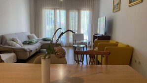 A living room with a sofa, a yellow couch, a dining table with a vase of flowers, a TV, and a window with curtains. - House Katy (Ploce)