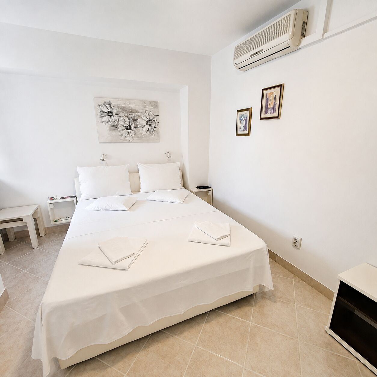 A hotel room with a bed, bedside tables, a wall-mounted air conditioner, and a painting.