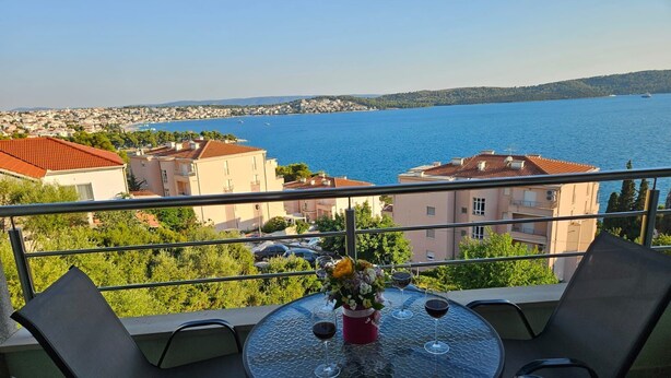 Ein Balkon mit rundem Tisch und Stühlen, mit Blick auf ein Gewässer und eine Küstenstadt.