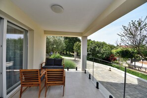 A balcony with a sofa and chairs, a glass door, and a view of a garden and trees. - Apartment Vergotin (Porec)