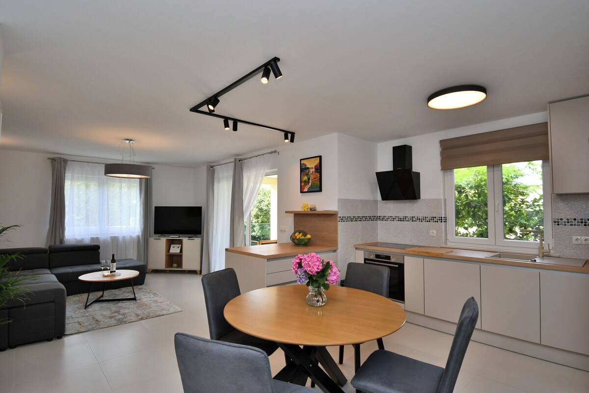 A modern living room with a sofa, coffee table, and dining table with chairs. There is a kitchen area with a sink, stove, and cabinets. A TV is mounted on the wall, and there are windows with curtains.