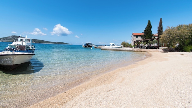 A beach with boats, clear water, and a building with a red roof. - Apartments Delfin (Vodice)