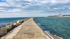 Ein Küstenweg mit einem Steg, der sich ins Meer erstreckt, bewölkter Himmel und ein Gebäude in der Ferne am Ufer.