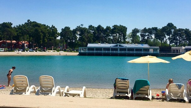 Ein Strand mit Liegestühlen und Sonnenschirmen, ein Boot anlegender am Wasser, und Gebäude im Hintergrund.