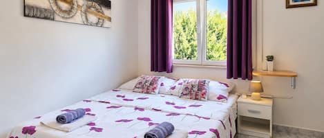 A bedroom with a bed, a nightstand, a lamp, a window with curtains, and a wall art piece.