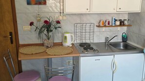 Eine kompakte Küche mit Kühlschrank, Spüle, Herd und einer kleinen Arbeitsfläche. Zwei Stühle mit Kissen, eine Vase mit Blumen und eine wandmontierte Steckdose.