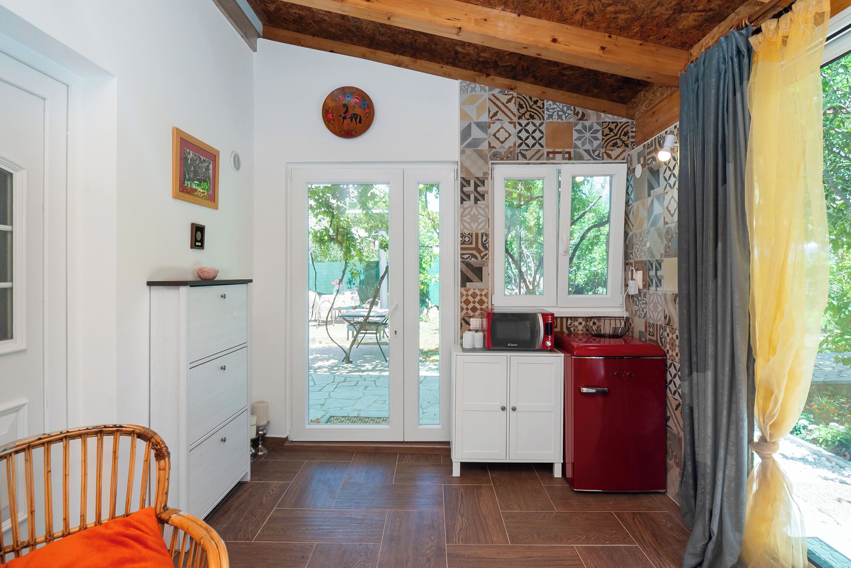 Quarto com teto de madeira, armários brancos, geladeira vermelha e vista para o exterior por uma porta de vidro.