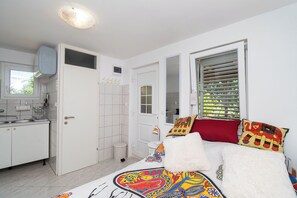 Ein Schlafzimmer mit Bett, Kissen, einem Fenster mit Jalousien, einer Tür und einem Badezimmer mit Waschbecken und Spiegel.