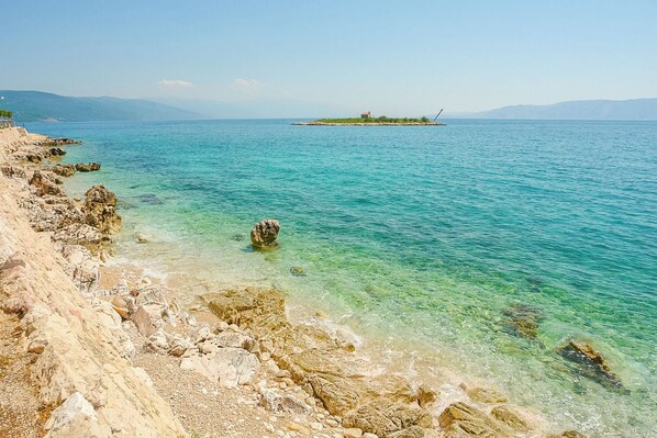 A coastal landscape with clear turquoise waters, rocky shoreline, and a distant island with a structure. - Apartments Marija (Novi Vinodolski)