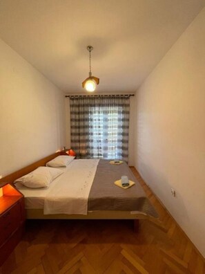Une petite chambre avec un lit simple, un plancher en bois, un lit recouvert de draps blancs, une table de chevet avec une lampe et une fenêtre ornée de rideaux à carreaux.