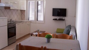 Ett kompakt kök med en spis, ugn och vita skåp. Där finns ett matbord med stolar och en liten krukväxt. En tv är monterad på väggen ovanför en soffa.