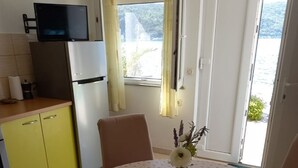 A dining area with a round table covered by a floral tablecloth, yellow cabinets, a television on a stand, and a view of the outdoors through a window with curtains. - Apartment More 1 (Savar)