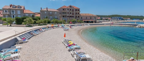 Une plage de galets avec des chaises longues et des parasols, une piscine à l’eau cristalline et des bâtiments en arrière-plan.