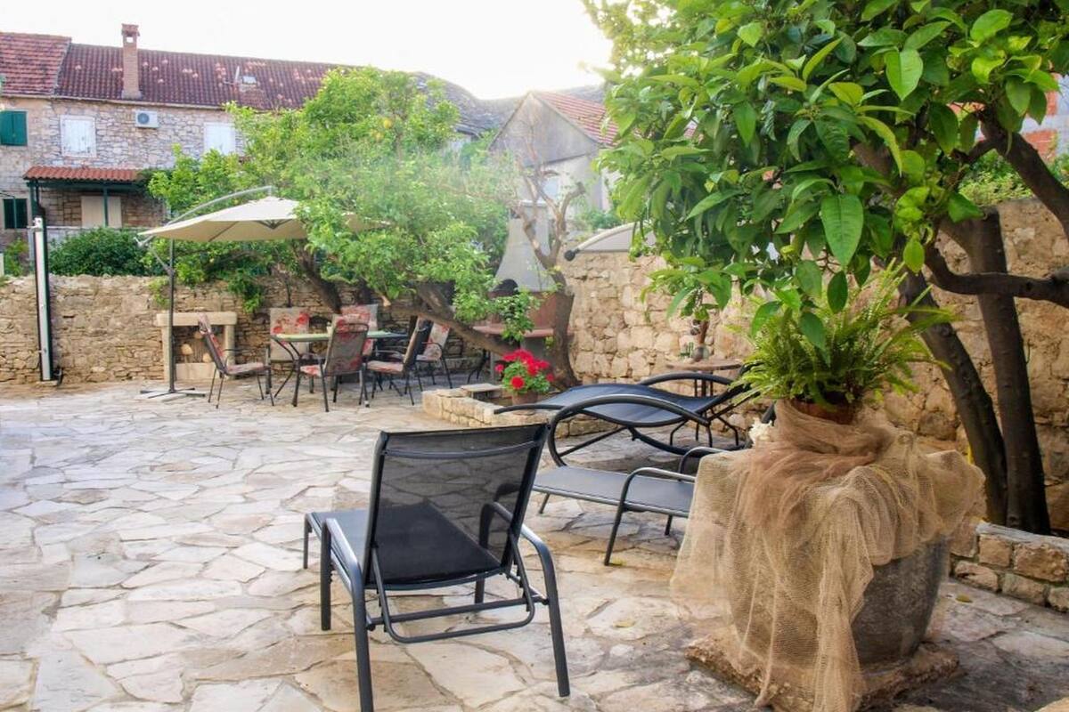 An outdoor patio with stone flooring, metal chairs, and tables. There is a large potted plant and a stone wall in the background.