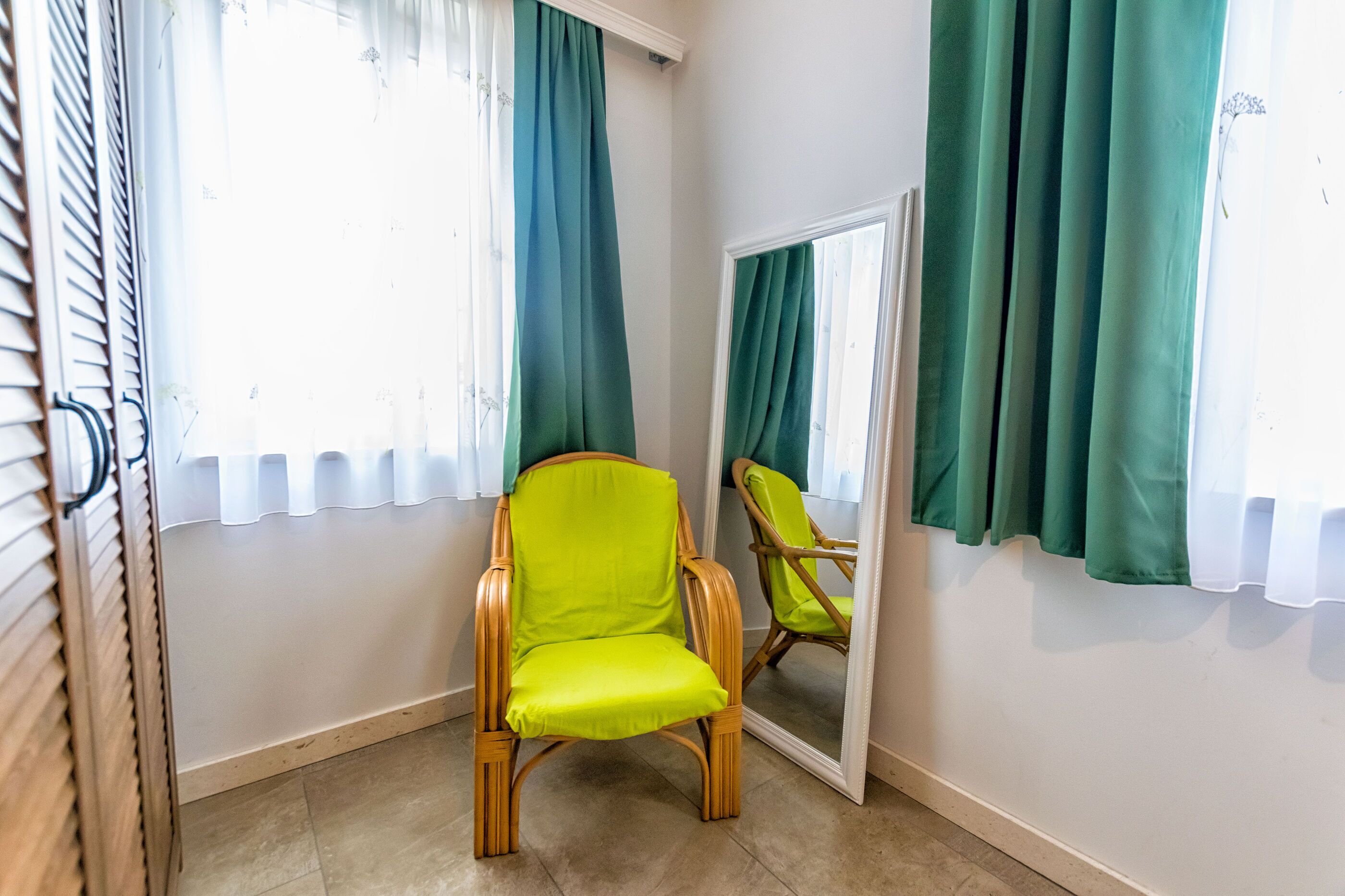 Ein Zimmer mit einem einzelnen grünen Stuhl, einem Spiegel und einem Fenster mit Vorhängen.