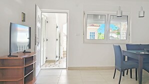 A modern living room with a flat-screen TV on a wooden stand, a dining area with a table and chairs, and a view through a door to an outdoor area with a tiled floor. - Apartments Helen (Rogoznica)