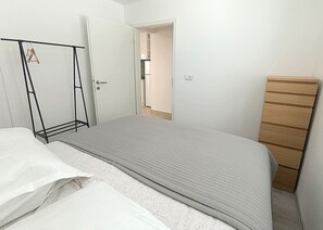 Une chambre à coucher comprenant un lit, une commode en bois, un portant à vêtements et une porte donnant sur une salle de bain.