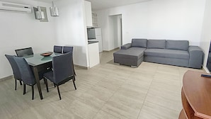 A modern living room with a dining area, grey sofa, and a small kitchen. - Apartments Helen (Rogoznica)