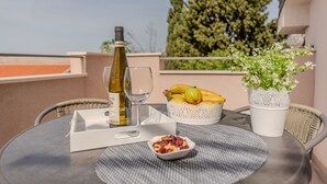 A table with a bottle of wine, a glass, a basket of fruit, and a small bowl of berries on a balcony with a potted plant and a view of trees. - Apartments Mil (Karlobag)