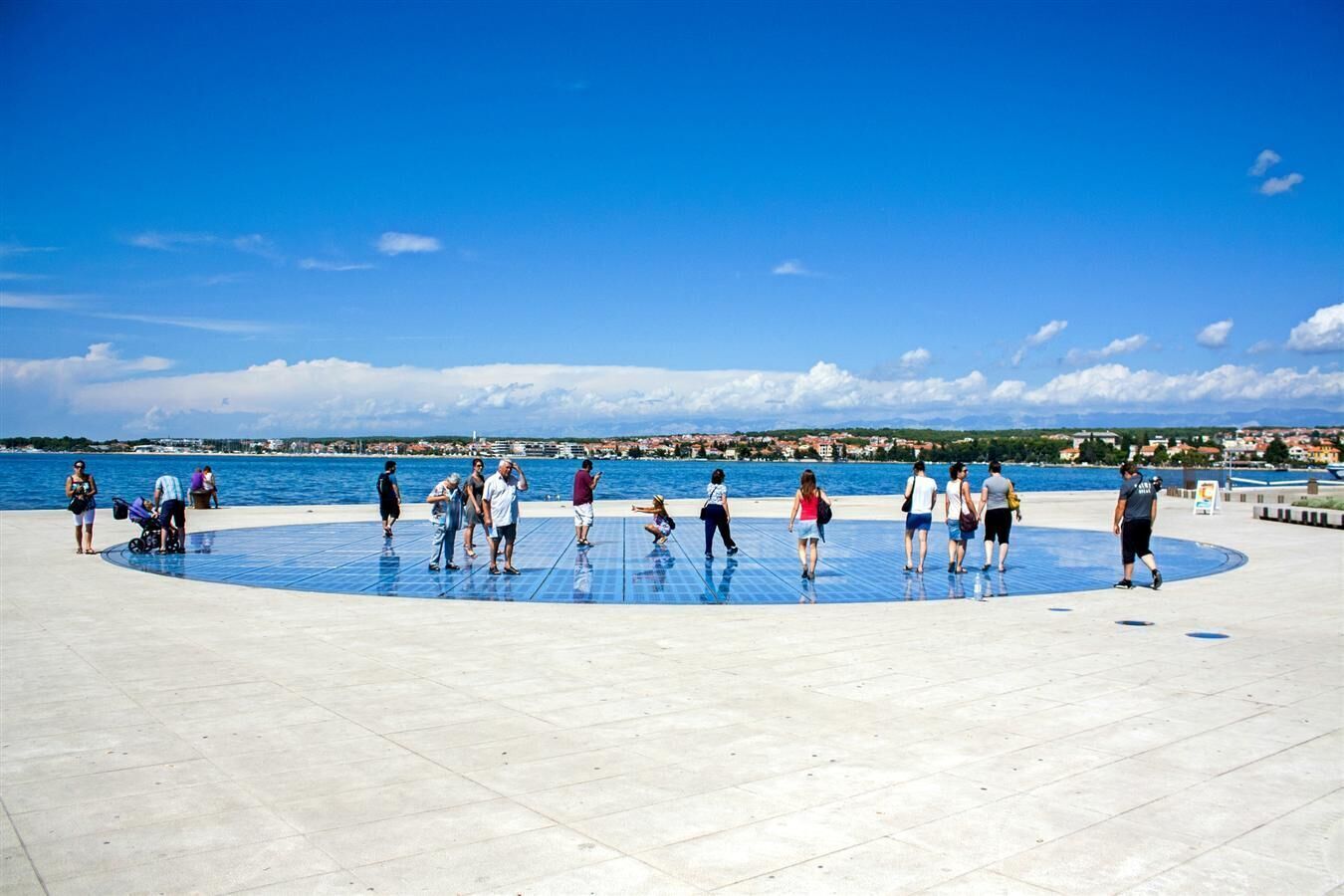 A public square with a circular blue floor, surrounded by a waterfront promenade.