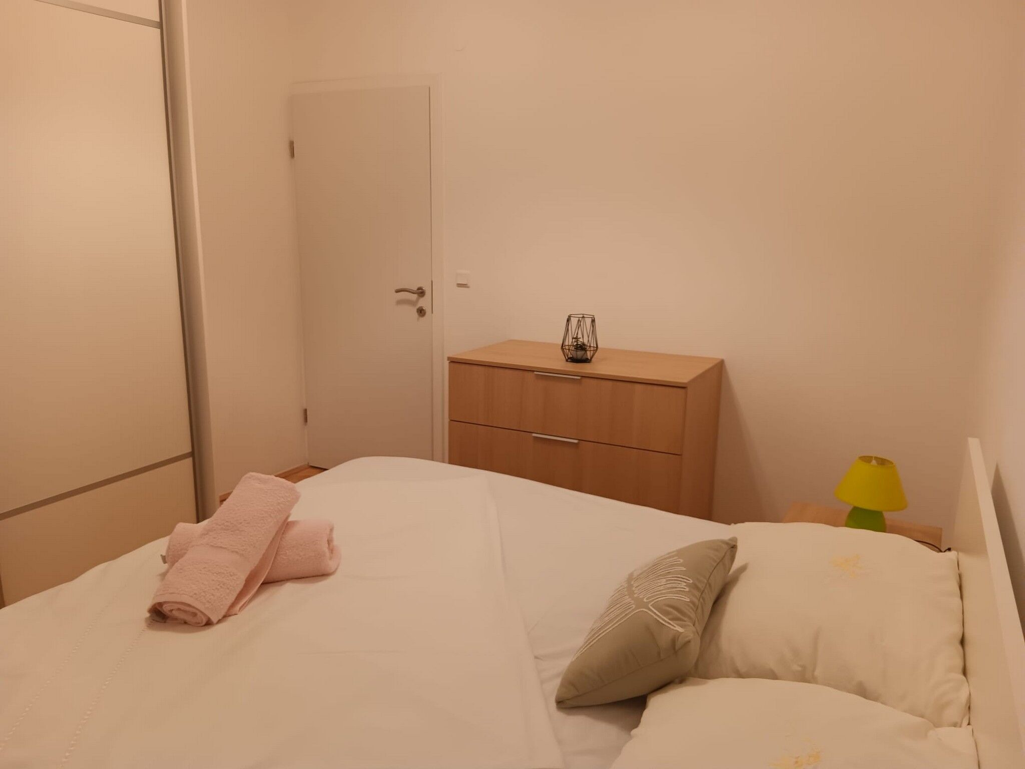 Cama bem arrumada com toalhas cor-de-rosa e um travesseiro decorativo, mesa de cabeceira de madeira com luminária e porta fechada.