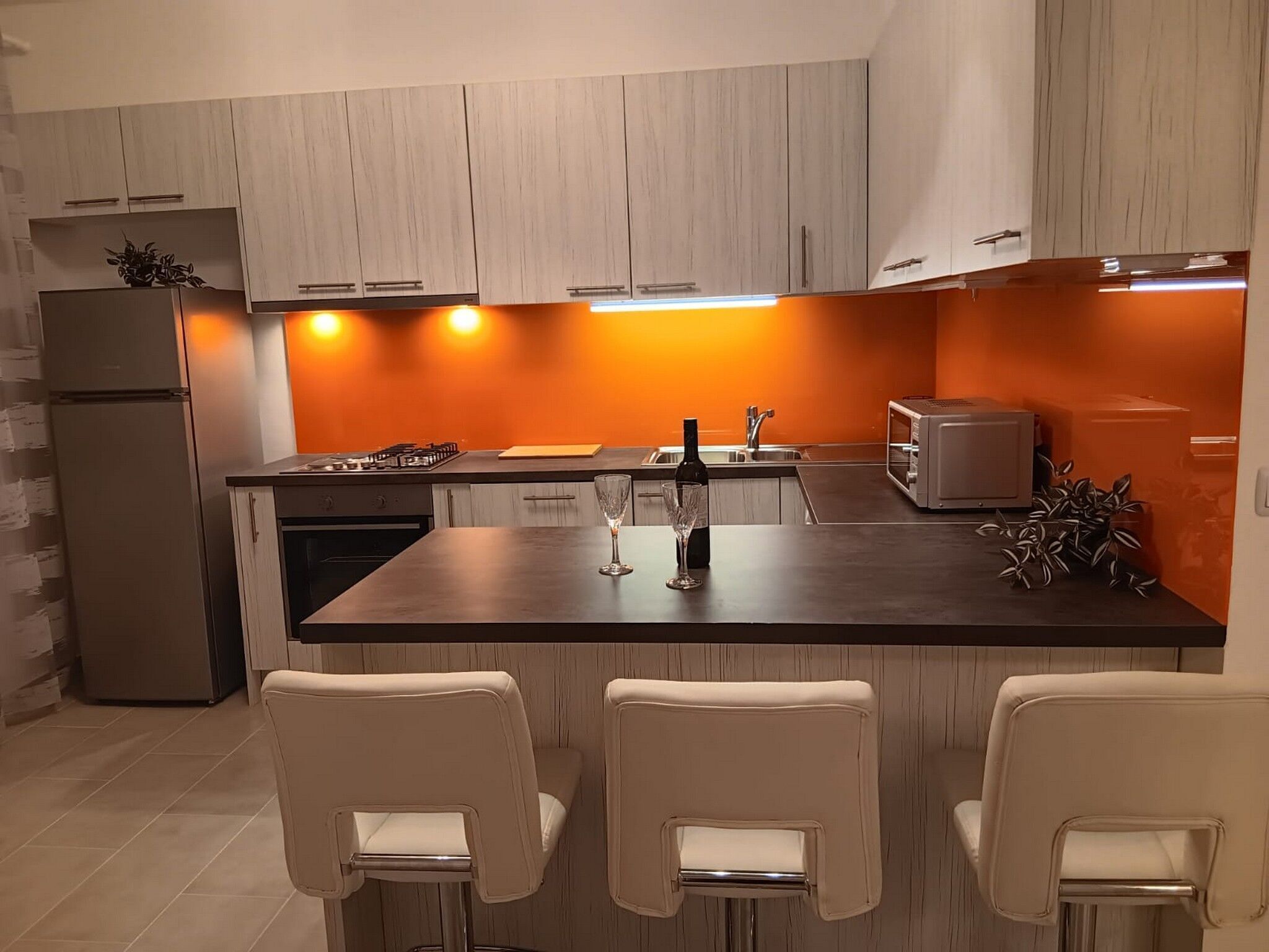 A modern kitchen with light wood cabinets, a central island with bar stools, a built-in oven, and a microwave.