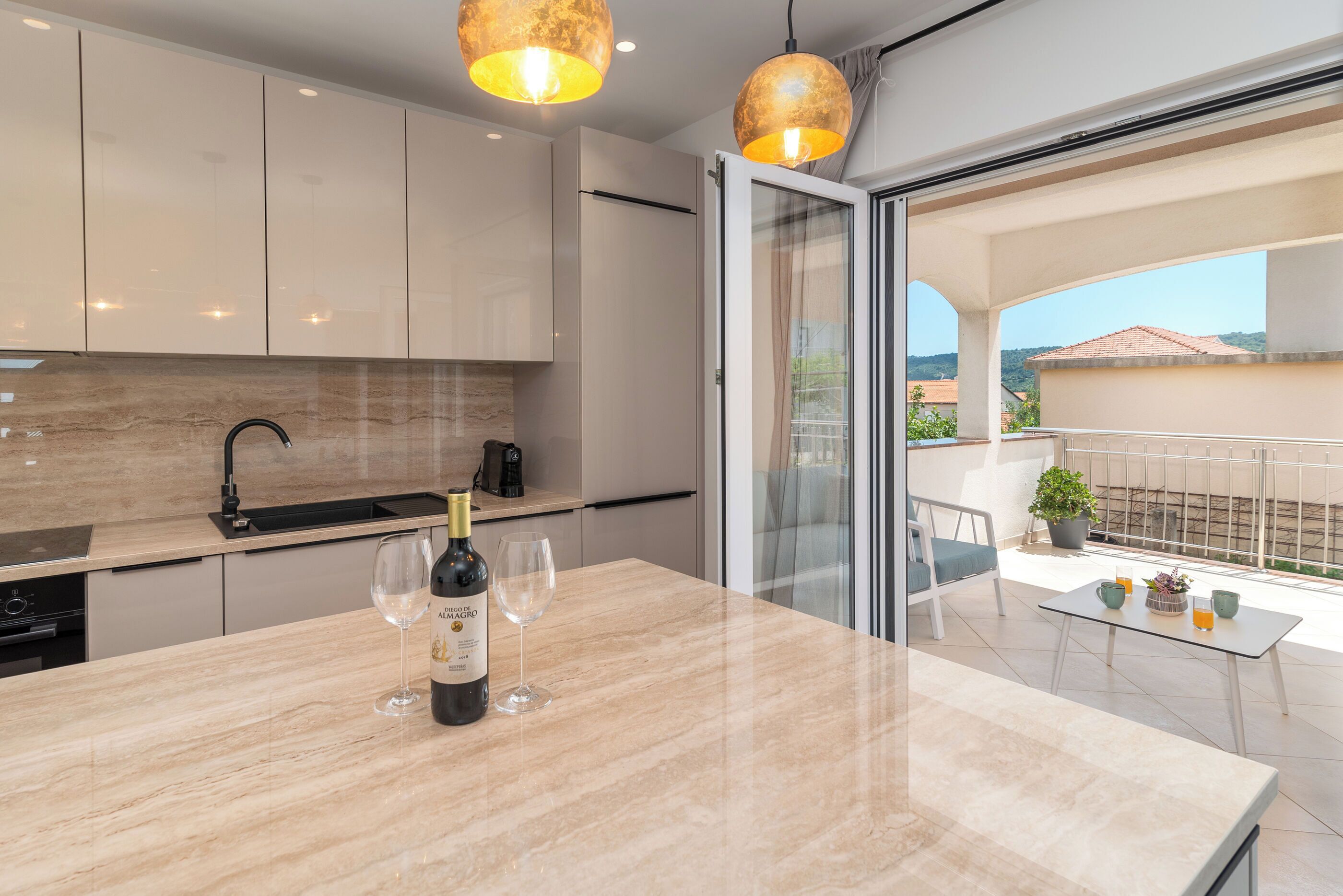 A modern kitchen with a marble countertop, wine bottle, and glasses. It features a sliding glass door leading to a balcony with a potted plant and a table.