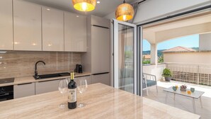 Una cocina moderna con encimera de mármol, una botella de vino y copas. Cuenta con una puerta corredera de cristal que da a un balcón con una planta en maceta y una mesa.