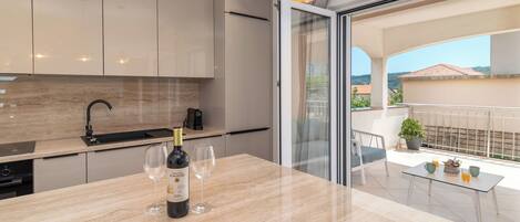 A modern kitchen with a marble countertop, wine bottle, and glasses. It features a sliding glass door leading to a balcony with a potted plant and a table.