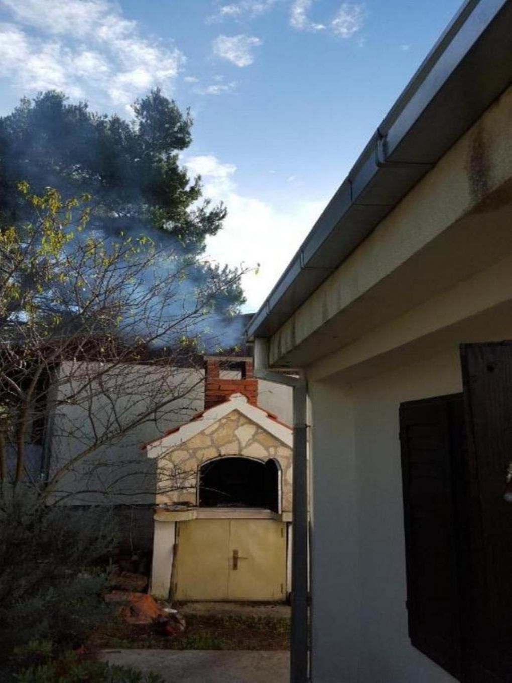 A stone-built structure with a chimney, possibly a fireplace, in a backyard with a tree and a closed garage door.
