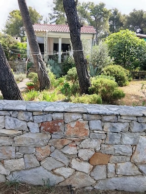 Eine Steinsichtmauer, ein Haus mit Ziegeldach und umgebende Grünflächen.