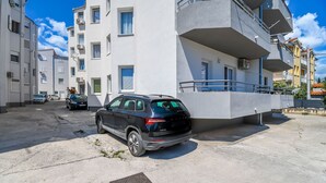 A black SUV parked in a concrete parking area with a white building featuring balconies and satellite dishes in the background. - Apartment Agi (Kastela)