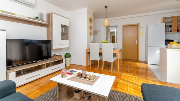 Egy modern nappali, melyben található egy síkképernyős televízió, egy étkező, ahol egy fa asztal és székek állnak, valamint egy konyha, melyben fehér szekrények és egy hűtőszekrény van.