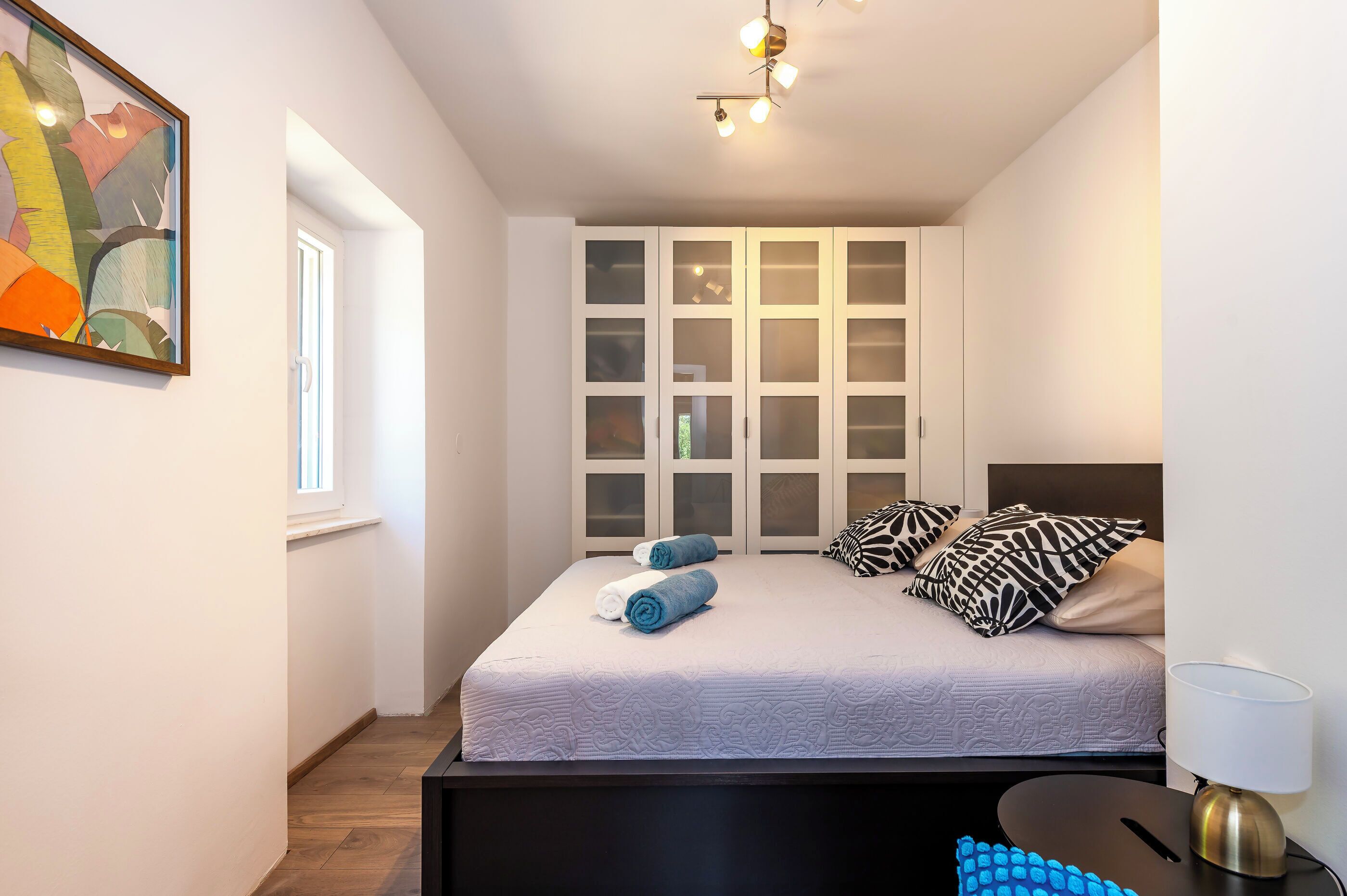 Ein Schlafzimmer mit einem Bett, einem Nachttisch, einer Lampe, einem Fenster und einem Wandbild.