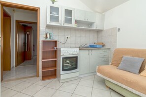 Eine kleine Küche mit Einbauofen, Spüle und weißen Schränken. Es gibt ein Holzregal und ein beiges Sofa mit Kissen.