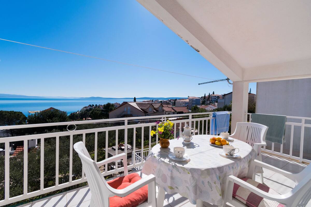 Un balcon avec une table blanche dressée pour le thé, un vase de fleurs et une vue sur la mer et les bâtiments.