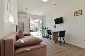 A modern living room with a beige sofa, a dining area with a table and chairs, a flat-screen TV mounted on the wall, and a glass door leading outside. - Apartments Kati (Seget)