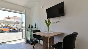 A modern room with a wooden table, two black chairs, a flat-screen TV mounted on the wall, and a large window offering a view of parked cars outside. - Apartments Kati (Seget)