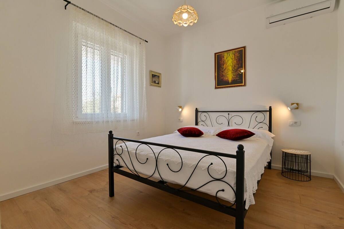 A bedroom with a metal bed frame, a window with sheer curtains, a wall-mounted light, and a small round table.