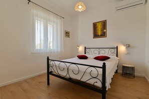 Un dormitorio con estructura de cama metálica, una ventana con cortinas translúcidas, una lámpara fijada a la pared y una mesita redonda pequeña.